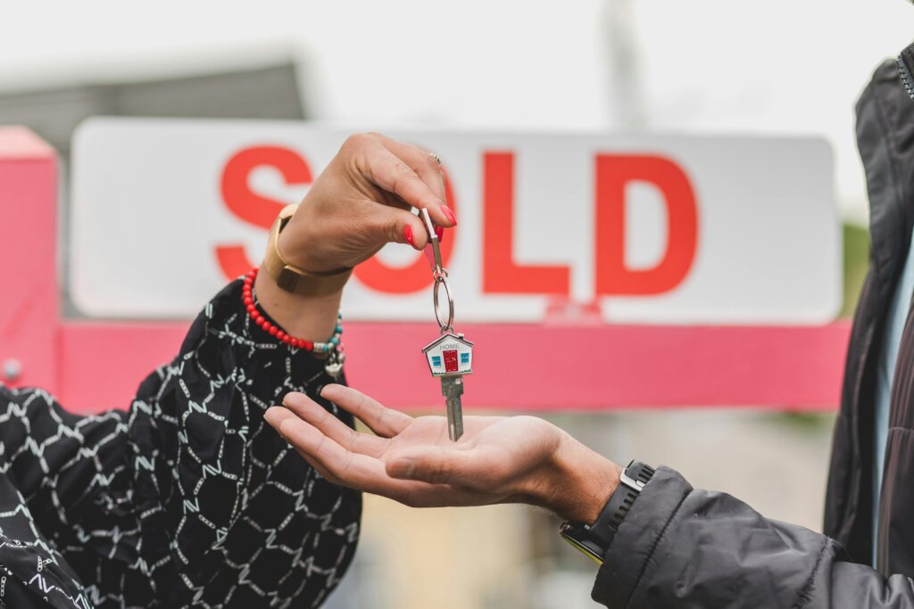 Person handing over house key in front of sold sign